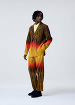 A person models HOMME PLISSÉ ISSEY MIYAKE's PAINT BRUSH CLOSE-UP: a striped yellow-to-red gradient suit with straight pants over a yellow shirt, paired with black shoes. They wear short dreadlocks and look slightly off to the side in a studio.