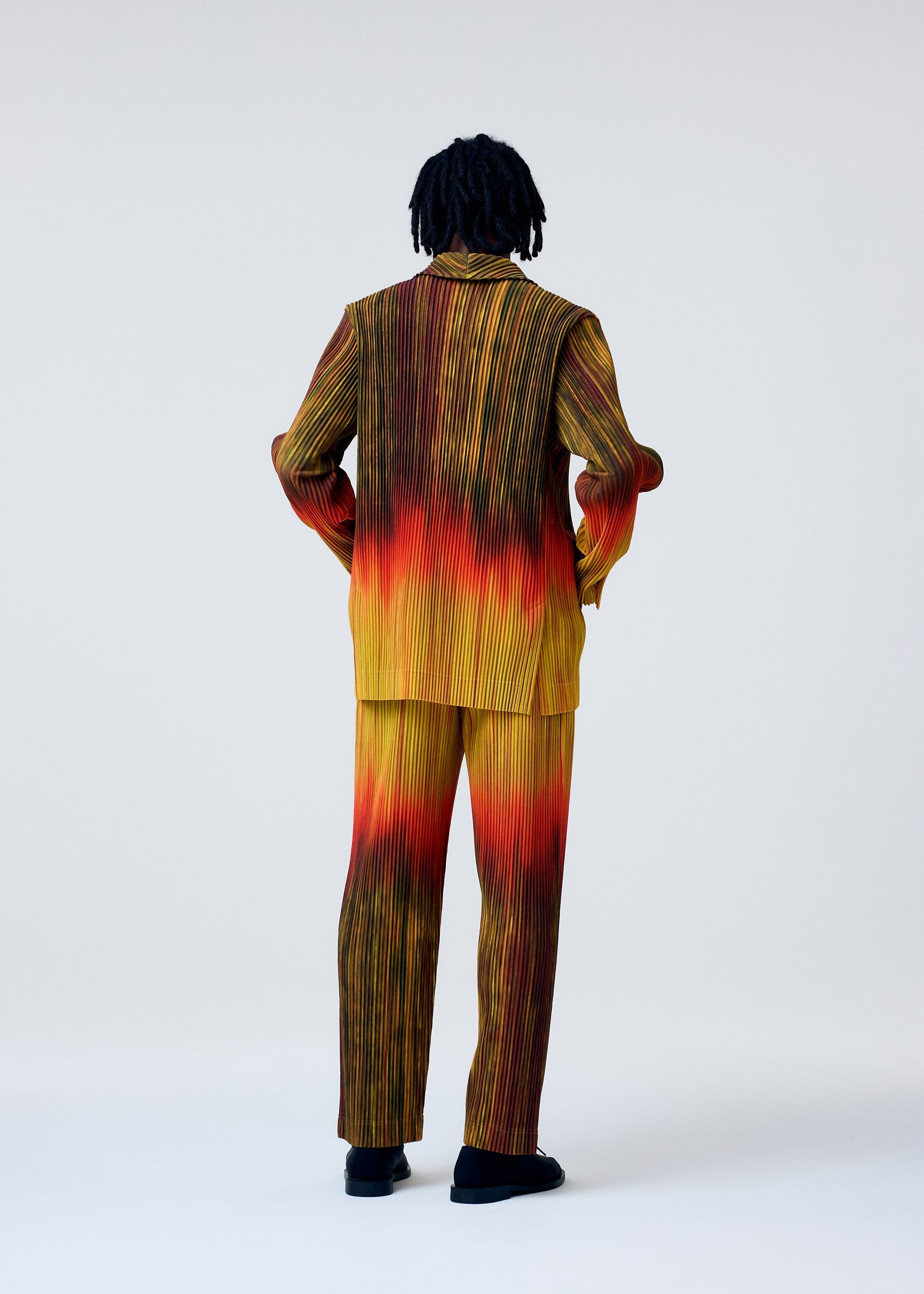 Zoomed view A person with short dreadlocks stands facing away, wearing the HOMME PLISSÉ ISSEY MIYAKE PAINT BRUSH CLOSE-UP pleated suit, featuring a gradient of yellow, red, and black, hands in pockets against a plain light background.