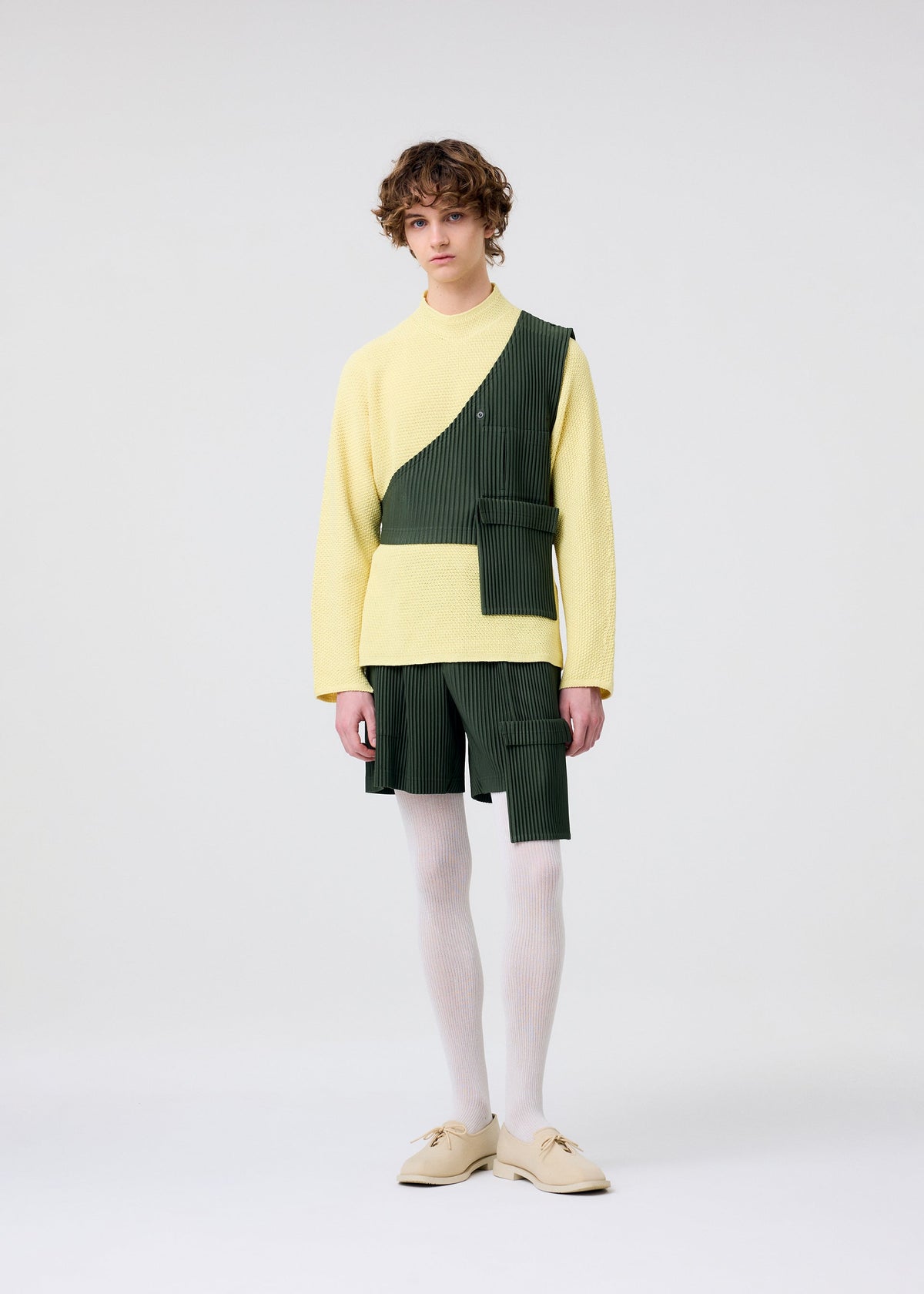 A person with short, curly brown hair stands against a plain background wearing HOMME PLISSÉ ISSEY MIYAKE PAINTER’S GEAR: a yellow sweater, green painter shorts with utility pockets and asymmetrical overlay and belt, white tights, and beige shoes. view 7 of 10