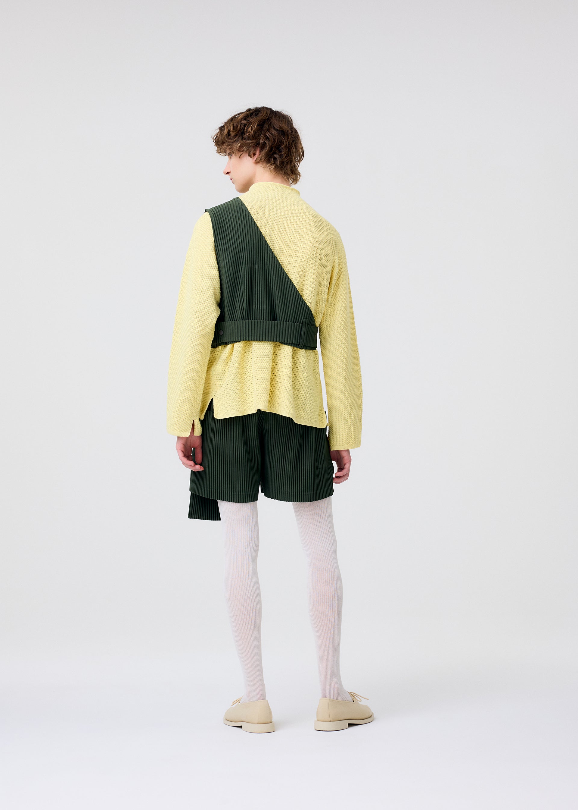 Zoomed view A person with curly hair, seen from behind, models HOMME PLISSÉ ISSEY MIYAKE’s PAINTER’S GEAR: a yellow sweater, green pleated painter shorts with utility pockets and sash, white tights, and cream shoes against a plain white background.