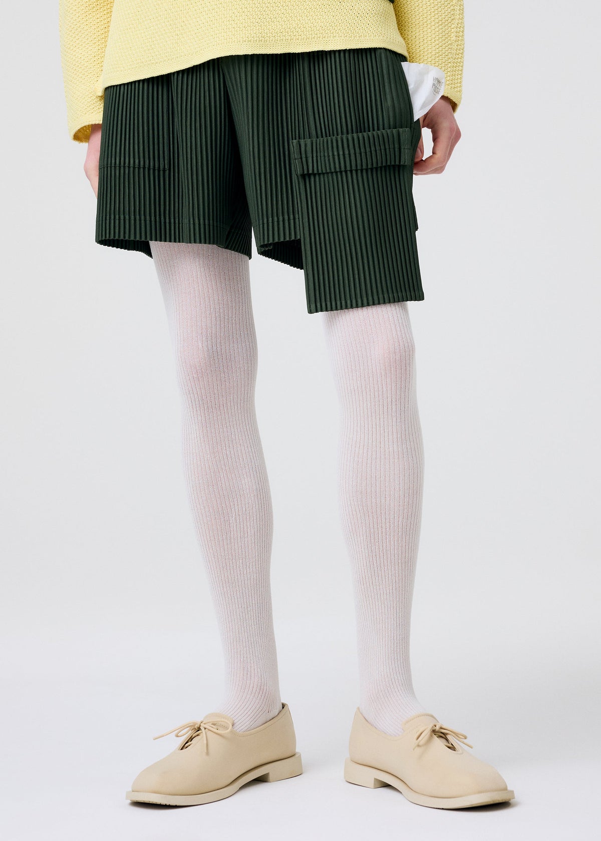 Model wears PAINTER’S GEAR painter shorts by HOMME PLISSÉ ISSEY MIYAKE, featuring utility and flap pockets. Styled with white ribbed tights, beige lace-up shoes, and a pale yellow textured sweater against a plain white background. view 9 of 10