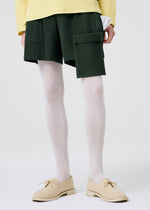 Model wears PAINTER’S GEAR painter shorts by HOMME PLISSÉ ISSEY MIYAKE, featuring utility and flap pockets. Styled with white ribbed tights, beige lace-up shoes, and a pale yellow textured sweater against a plain white background.
