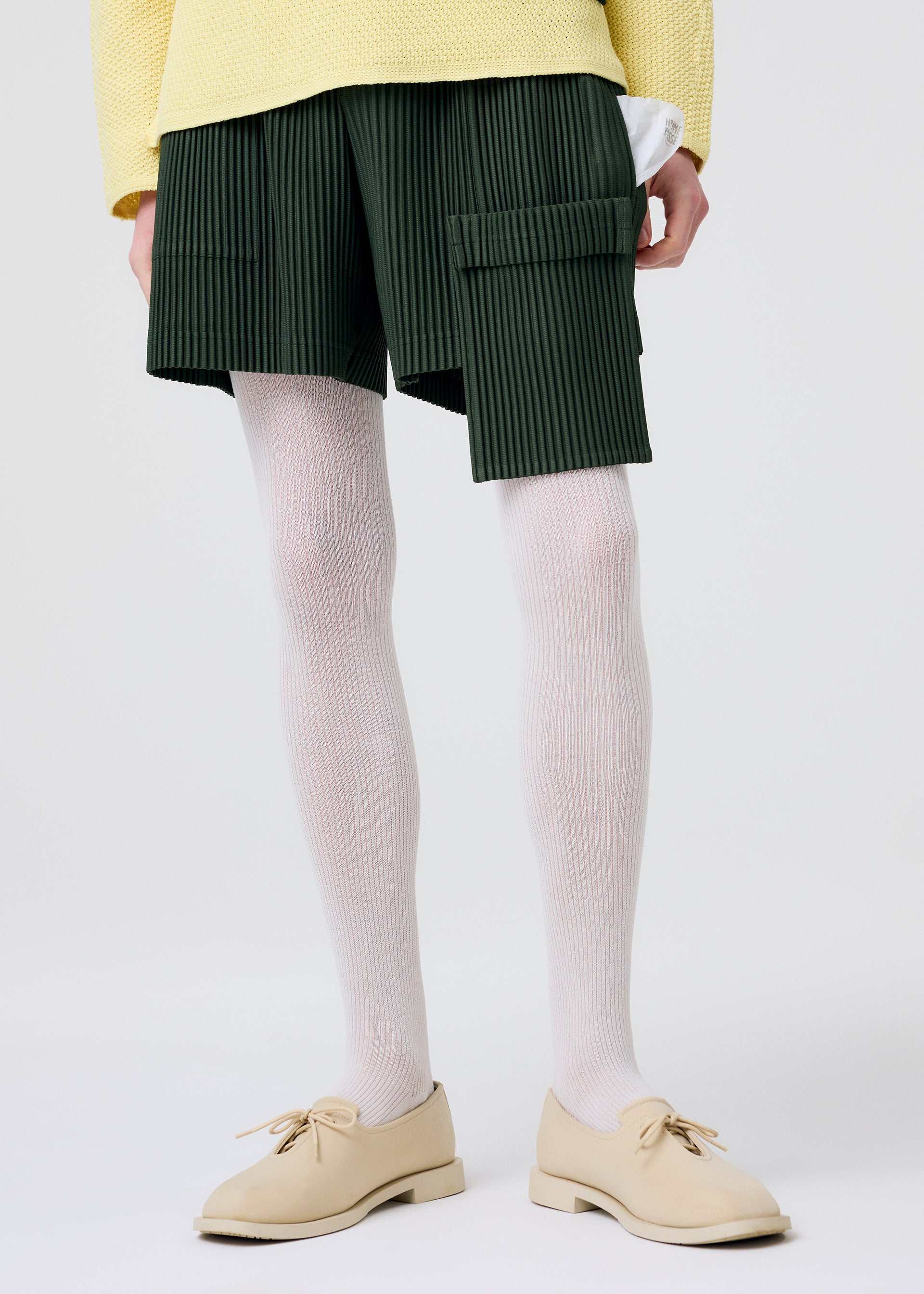 Zoomed view Model wears PAINTER’S GEAR painter shorts by HOMME PLISSÉ ISSEY MIYAKE, featuring utility and flap pockets. Styled with white ribbed tights, beige lace-up shoes, and a pale yellow textured sweater against a plain white background.