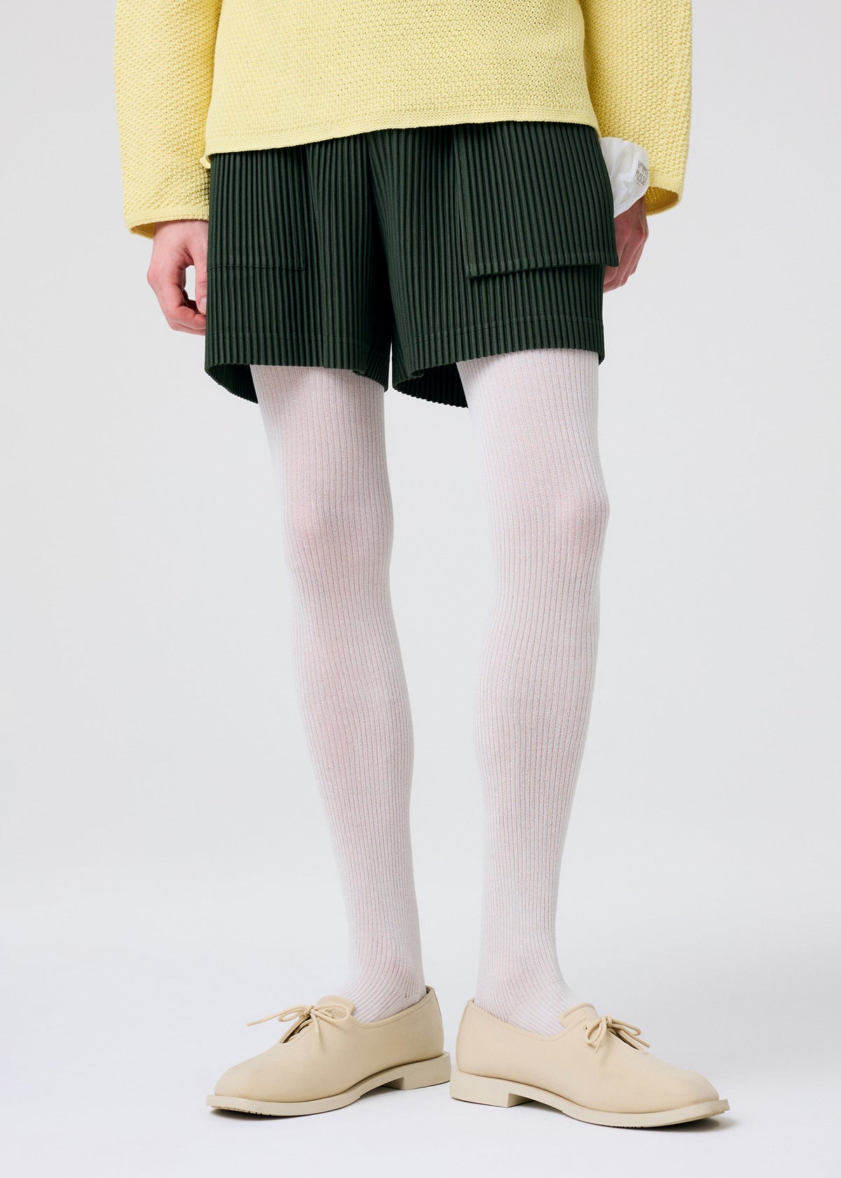 A person wearing HOMME PLISSÉ ISSEY MIYAKE’s PAINTER’S GEAR dark green shorts with utility pockets, paired with a yellow textured sweater, white ribbed tights, and beige lace-up shoes stands against a plain light background. view 10 of 10