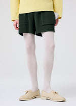 A person wearing HOMME PLISSÉ ISSEY MIYAKE’s PAINTER’S GEAR dark green shorts with utility pockets, paired with a yellow textured sweater, white ribbed tights, and beige lace-up shoes stands against a plain light background.
