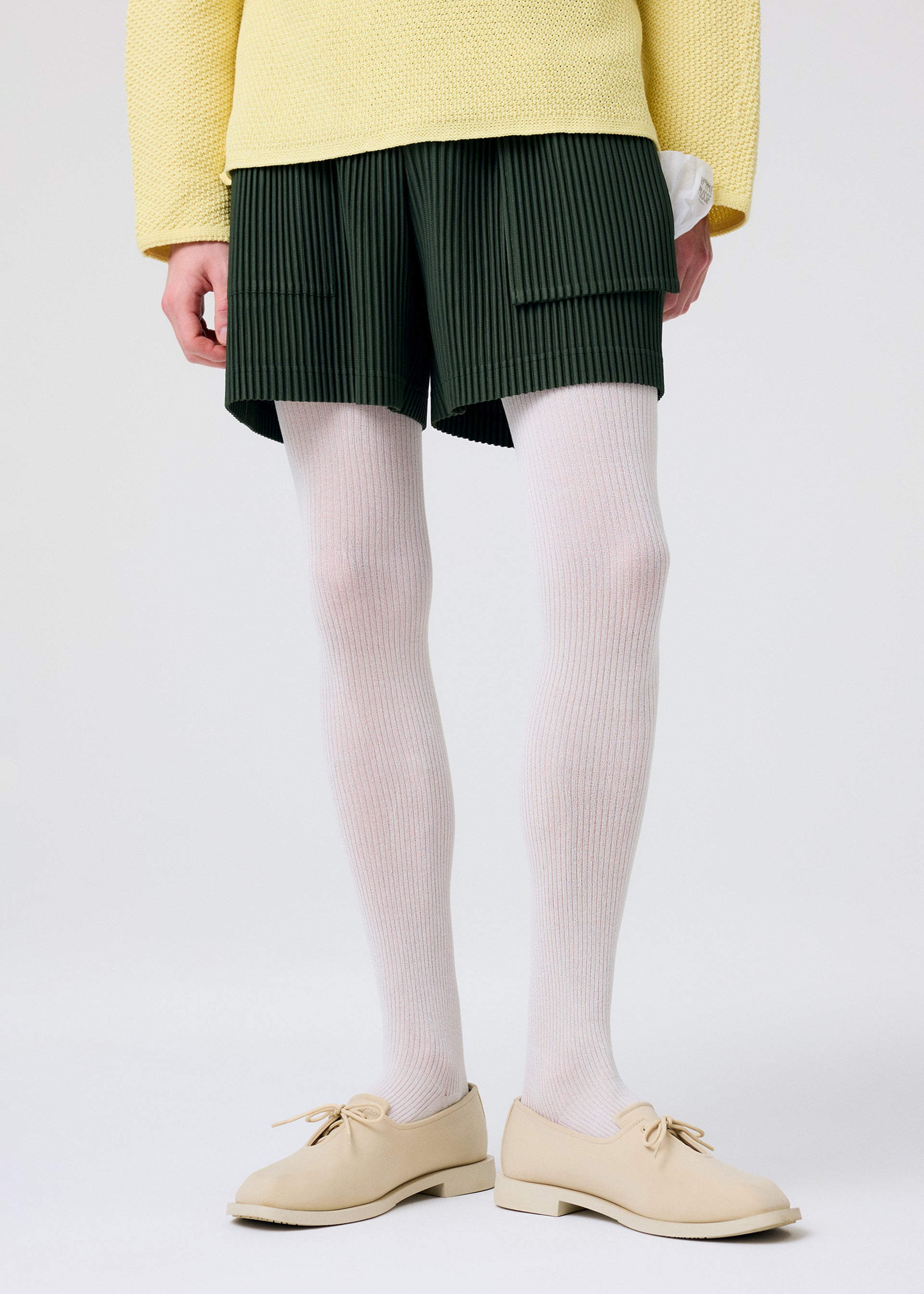 Zoomed view A person wearing HOMME PLISSÉ ISSEY MIYAKE’s PAINTER’S GEAR dark green shorts with utility pockets, paired with a yellow textured sweater, white ribbed tights, and beige lace-up shoes stands against a plain light background.