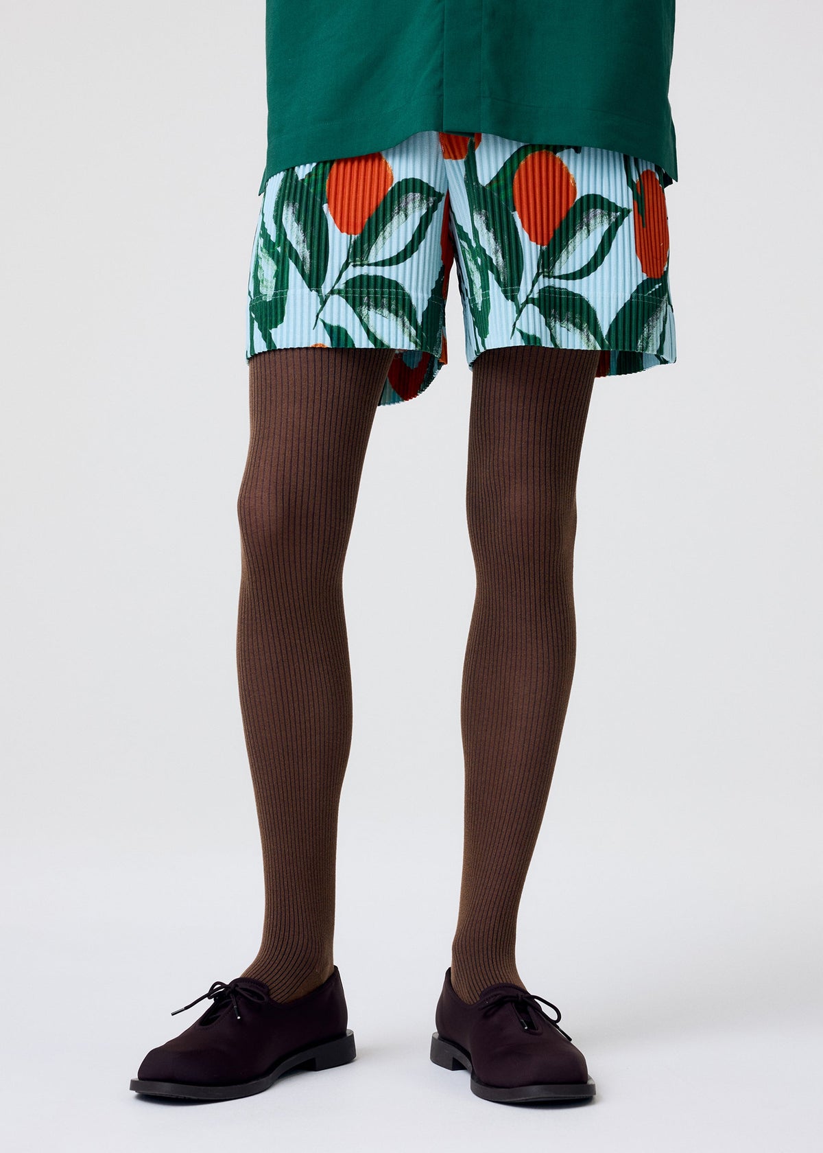 A person stands against a plain white background, wearing HOMME PLISSÉ ISSEY MIYAKE FRUITS & VEGETABLES light blue shorts featuring a green and orange fruit and leaf motif, paired with brown ribbed tights and black lace-up shoes. view 8 of 8