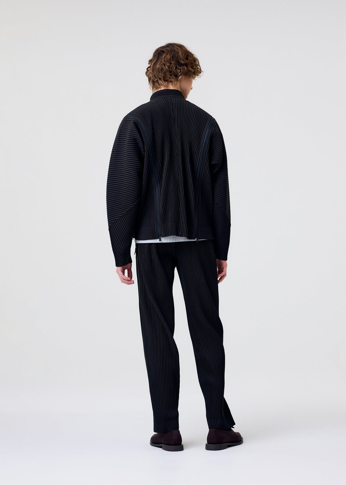 Facing away, a person with curly hair wears the HOMME PLISSÉ ISSEY MIYAKE MULTI-FASTENED—a loose, black pinstriped set with long sleeves and drawstring pants—paired with dark shoes against a light background. view 6 of 9