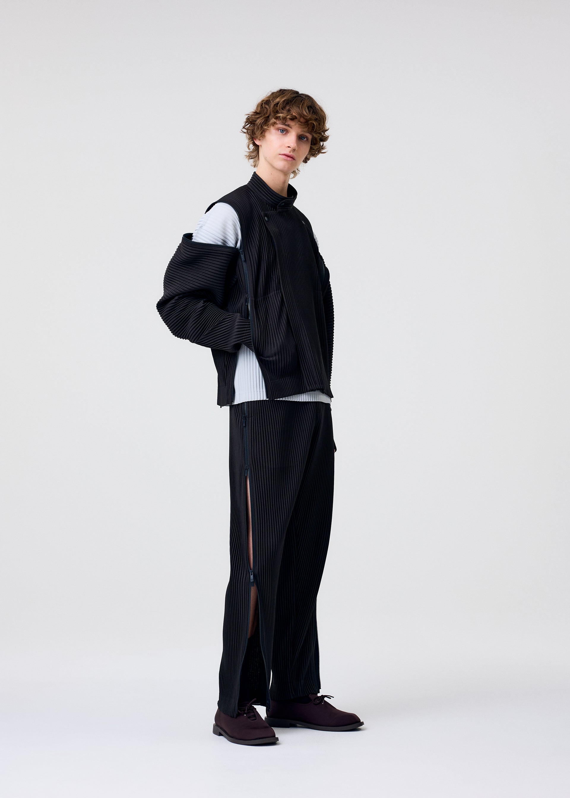 Zoomed view A person with curly hair stands against a plain background wearing the HOMME PLISSÉ ISSEY MIYAKE MULTI-FASTENED black pinstriped jacket draped over one shoulder and matching drawstring pants with side slits, a light gray top, and dark shoes.