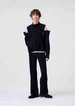 A person with curly hair stands against a plain light background, wearing HOMME PLISSÉ ISSEY MIYAKE’s MULTI-FASTENED black ribbed asymmetrical jacket, stylish drawstring pants, a light grey shirt, and dark shoes.