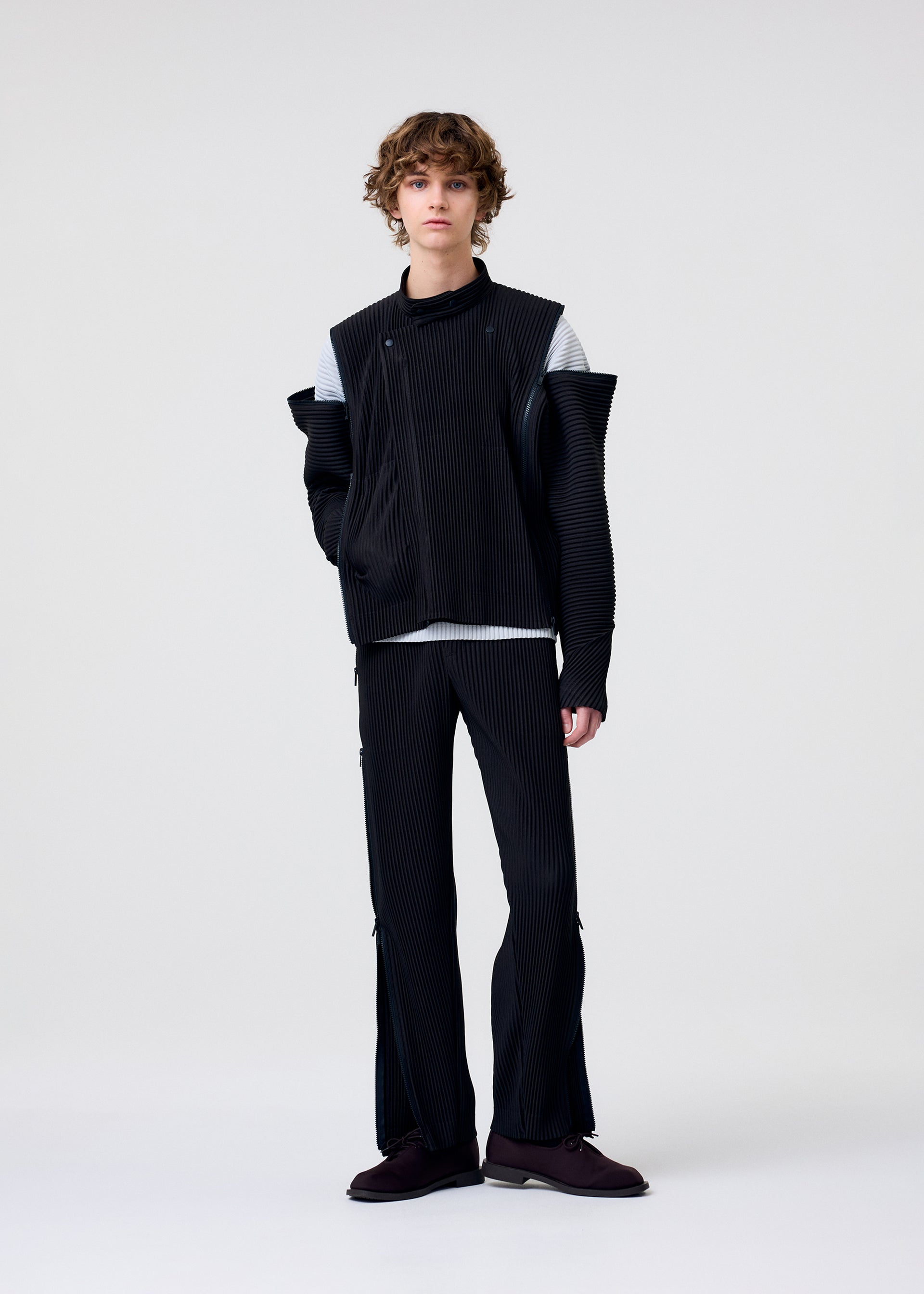 Zoomed view A person with curly hair stands against a plain light background, wearing HOMME PLISSÉ ISSEY MIYAKE’s MULTI-FASTENED black ribbed asymmetrical jacket, stylish drawstring pants, a light grey shirt, and dark shoes.