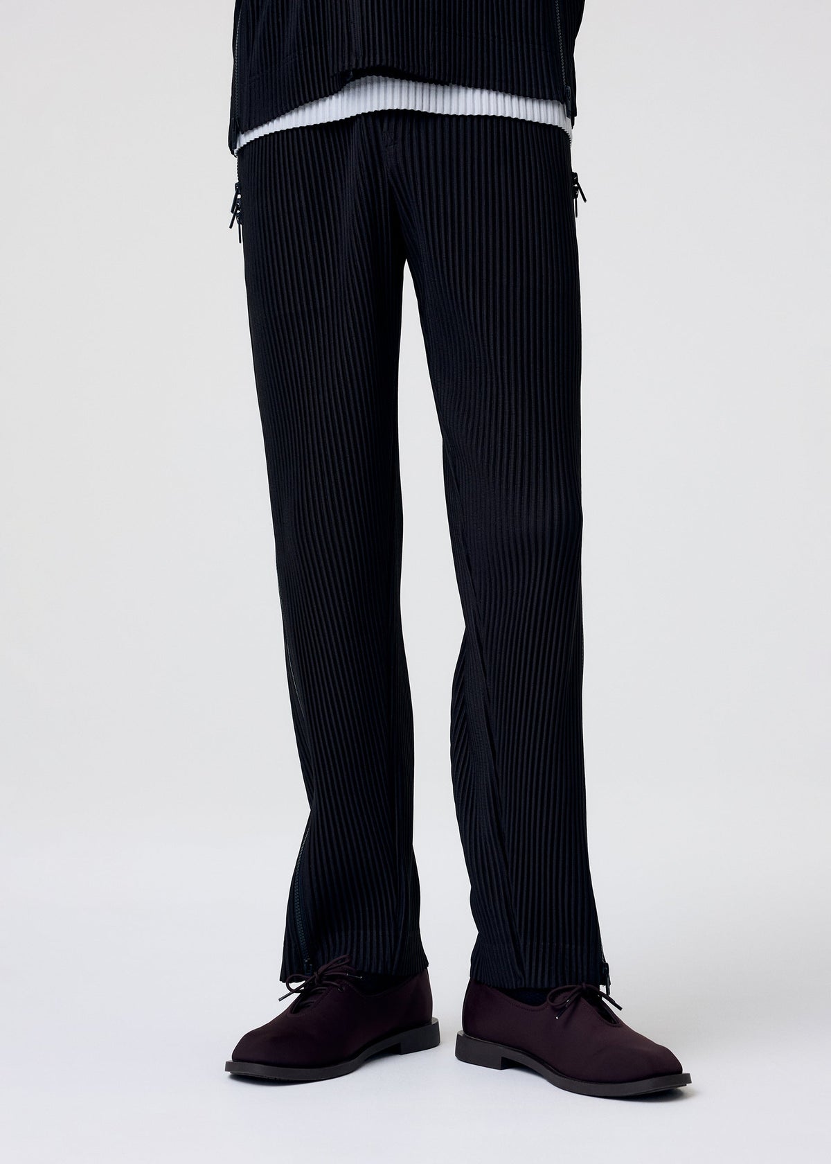 A person models the MULTI-FASTENED drawstring pants by HOMME PLISSÉ ISSEY MIYAKE in black pinstripes, paired with a white shirt and dark burgundy shoes; set against a plain white background with their upper body out of frame. view 9 of 9
