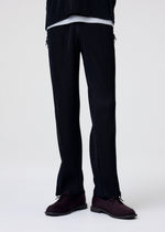 A person models the MULTI-FASTENED drawstring pants by HOMME PLISSÉ ISSEY MIYAKE in black pinstripes, paired with a white shirt and dark burgundy shoes; set against a plain white background with their upper body out of frame.