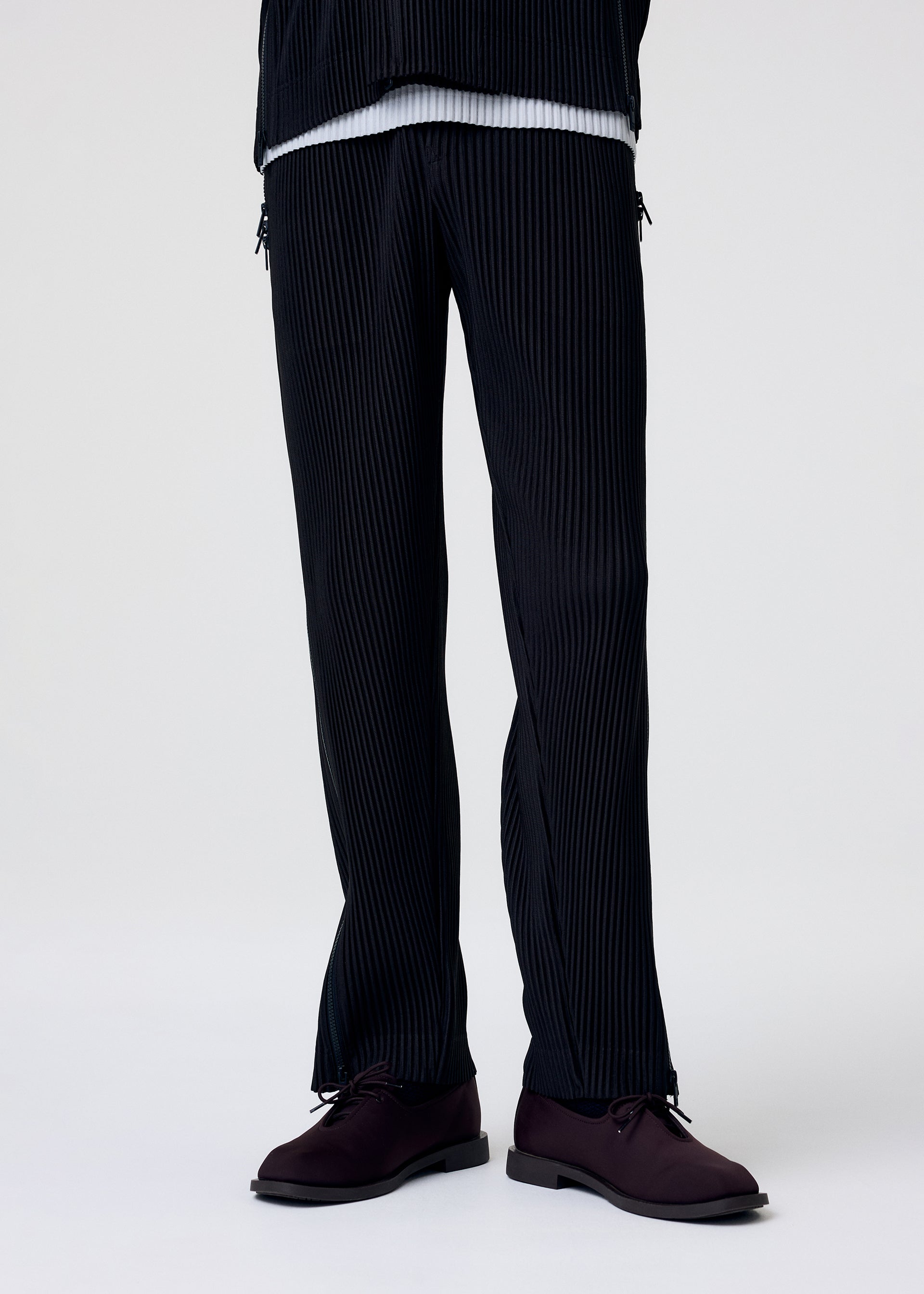 Zoomed view A person models the MULTI-FASTENED drawstring pants by HOMME PLISSÉ ISSEY MIYAKE in black pinstripes, paired with a white shirt and dark burgundy shoes; set against a plain white background with their upper body out of frame.