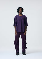 A person with medium-length, black, twisted hair faces forward, wearing a loose purple shirt and HOMME PLISSÉ ISSEY MIYAKE PLEATS BOTTOMS 1 pants with black shoes, set against a plain light background.