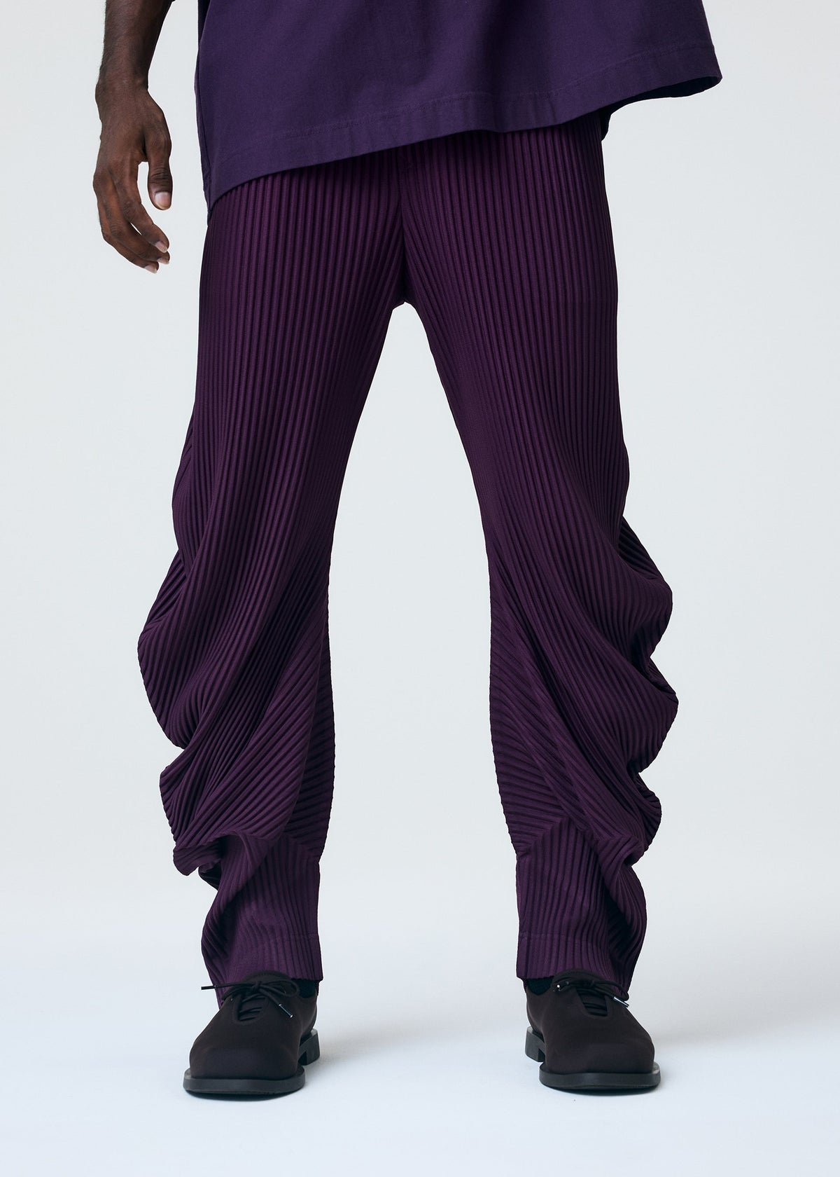 A person showcases HOMME PLISSÉ ISSEY MIYAKE's PLEATS BOTTOMS 1—loose, dark purple pleated ankle pants—paired with black shoes and a matching top, photographed from the waist down against a plain background. view 6 of 6