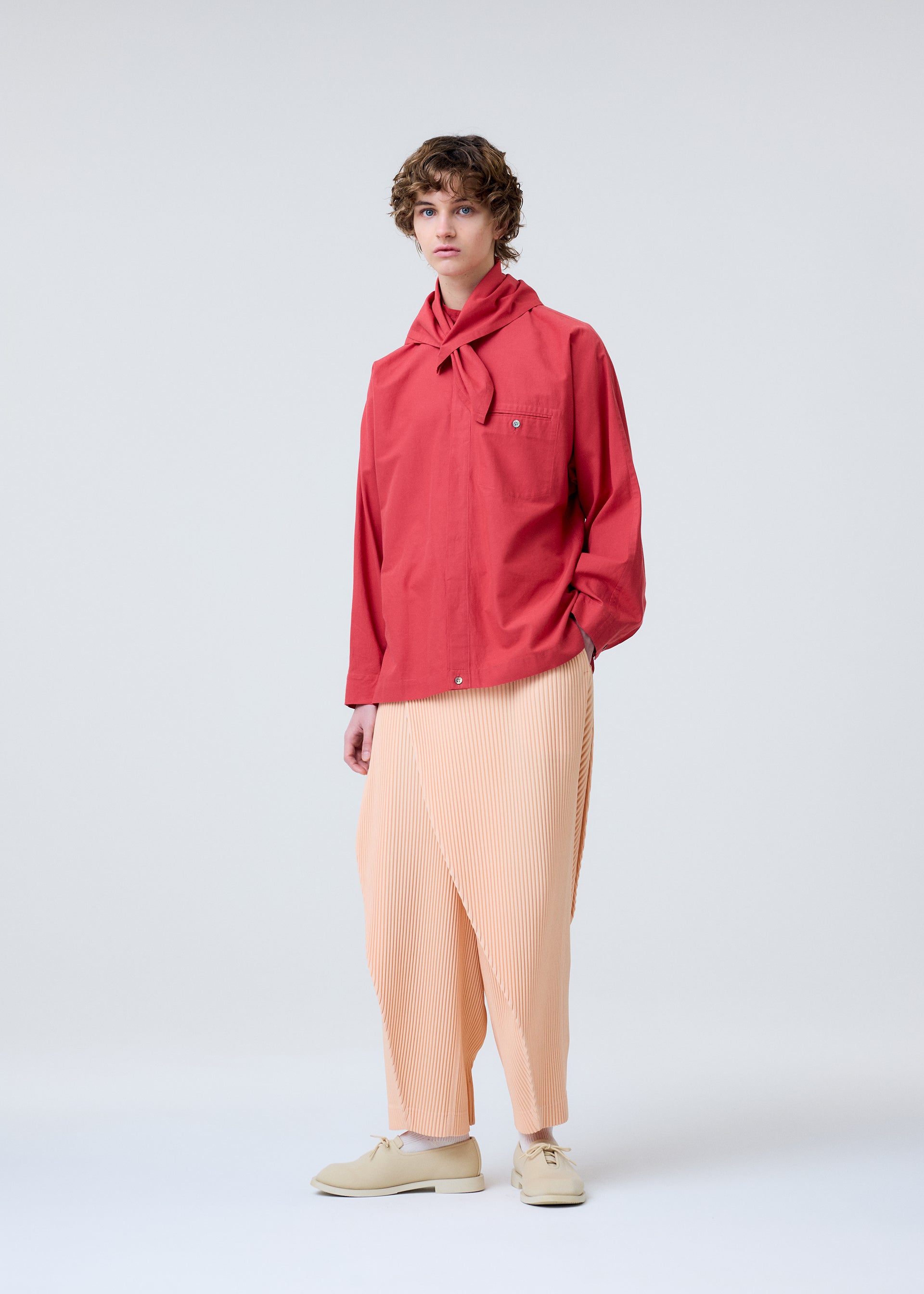 Zoomed view A person with short curly hair wears a bright red shirt with a tied collar and HOMME PLISSÉ ISSEY MIYAKE's PLEATS BOTTOMS 1, peach-colored and pleated. They stand against a plain light background in light shoes, showcasing elegant draping.