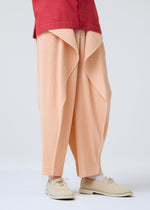A person models HOMME PLISSÉ ISSEY MIYAKE PLEATS BOTTOMS 1—loose, peach-toned wrap pants with elegant draping—paired with a red top, white socks, and cream shoes against a plain light background. Photo cropped above the waist.