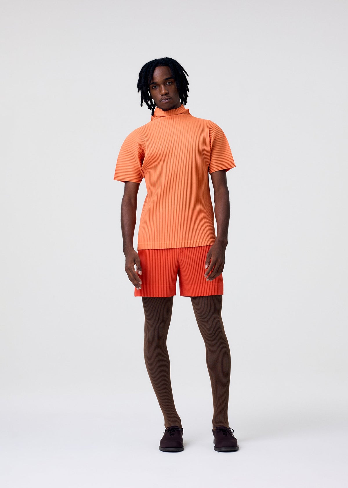 A person stands against a plain light background, wearing HOMME PLISSÉ ISSEY MIYAKE COLORFUL PLEATS BOTTOMS with a matching short-sleeve top, brown tights, dark shoes, and has short black hair and a neutral expression. view 8 of 8