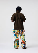A person stands facing away, wearing the HOMME PLISSÉ ISSEY MIYAKE PALETTE MARBLED pants—wide, handcrafted, with vibrant abstract patterns inspired by Kyoto marbling—and a loose dark brown ribbed shirt. The background is plain and light.