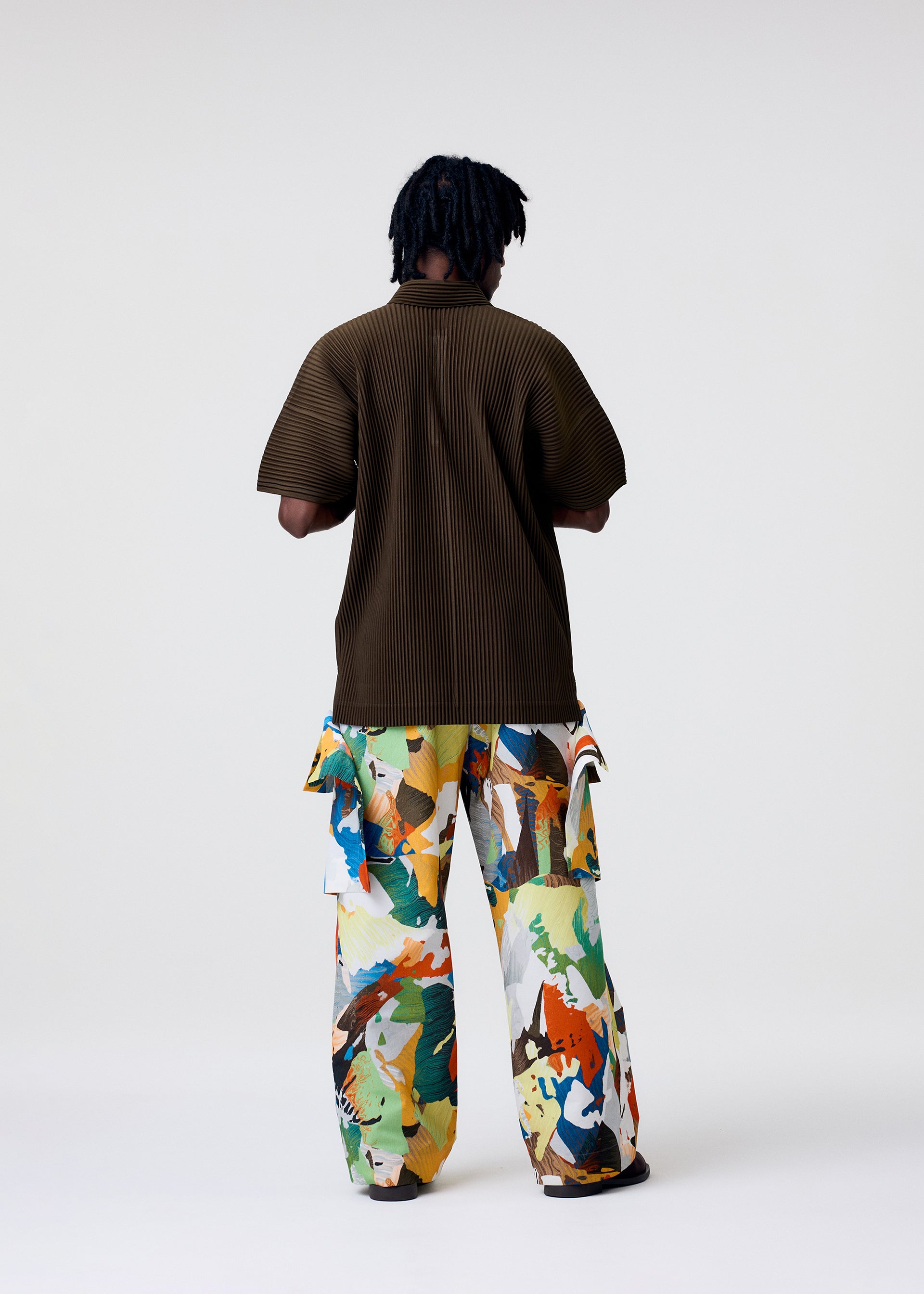 Zoomed view A person stands facing away, wearing the HOMME PLISSÉ ISSEY MIYAKE PALETTE MARBLED pants—wide, handcrafted, with vibrant abstract patterns inspired by Kyoto marbling—and a loose dark brown ribbed shirt. The background is plain and light.