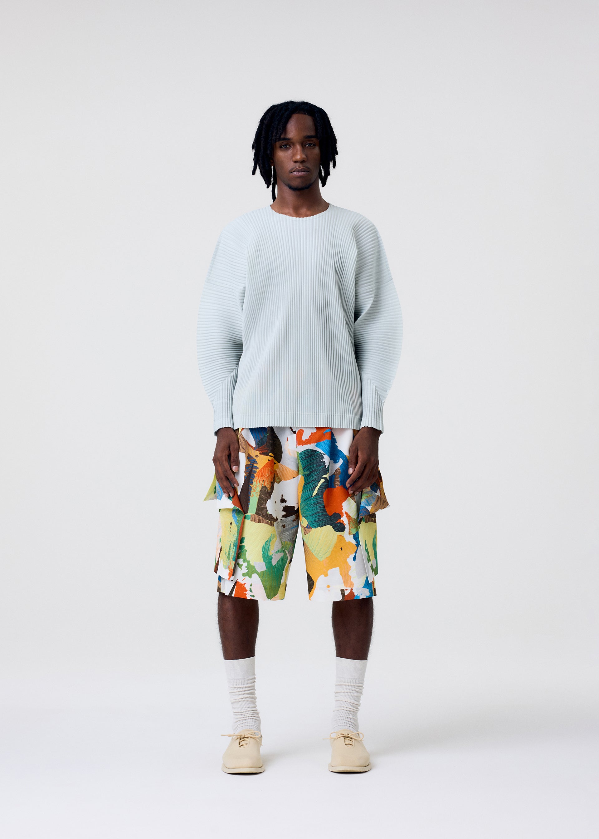 Zoomed view A person stands against a plain white background wearing a light ribbed sweater, HOMME PLISSÉ ISSEY MIYAKE PALETTE MARBLED shorts with a unique pattern, cream socks, and beige shoes.
