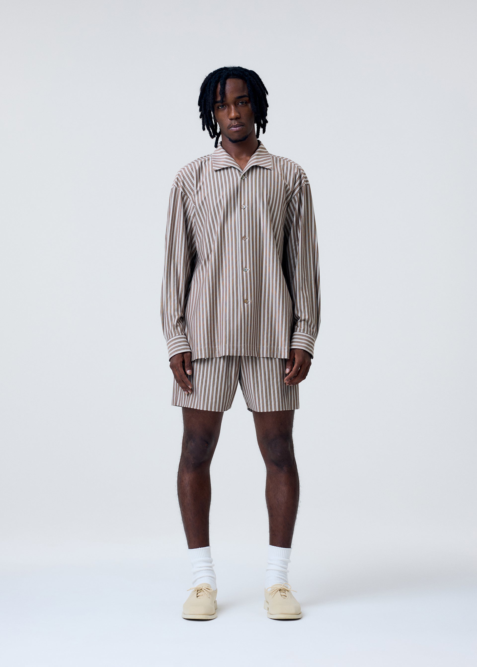 Zoomed view A person stands facing forward, wearing the STRIPE JERSEY set by HOMME PLISSÉ ISSEY MIYAKE—an Italian-inspired beige and brown striped button-up shirt with matching shorts—paired with white socks and cream shoes, set against a plain light background.