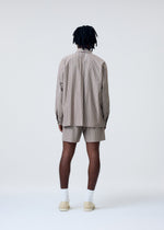 A person with dark, shoulder-length hair faces away, wearing the HOMME PLISSÉ ISSEY MIYAKE STRIPE JERSEY set in beige and brown, paired with white socks and beige shoes, against a plain light background.