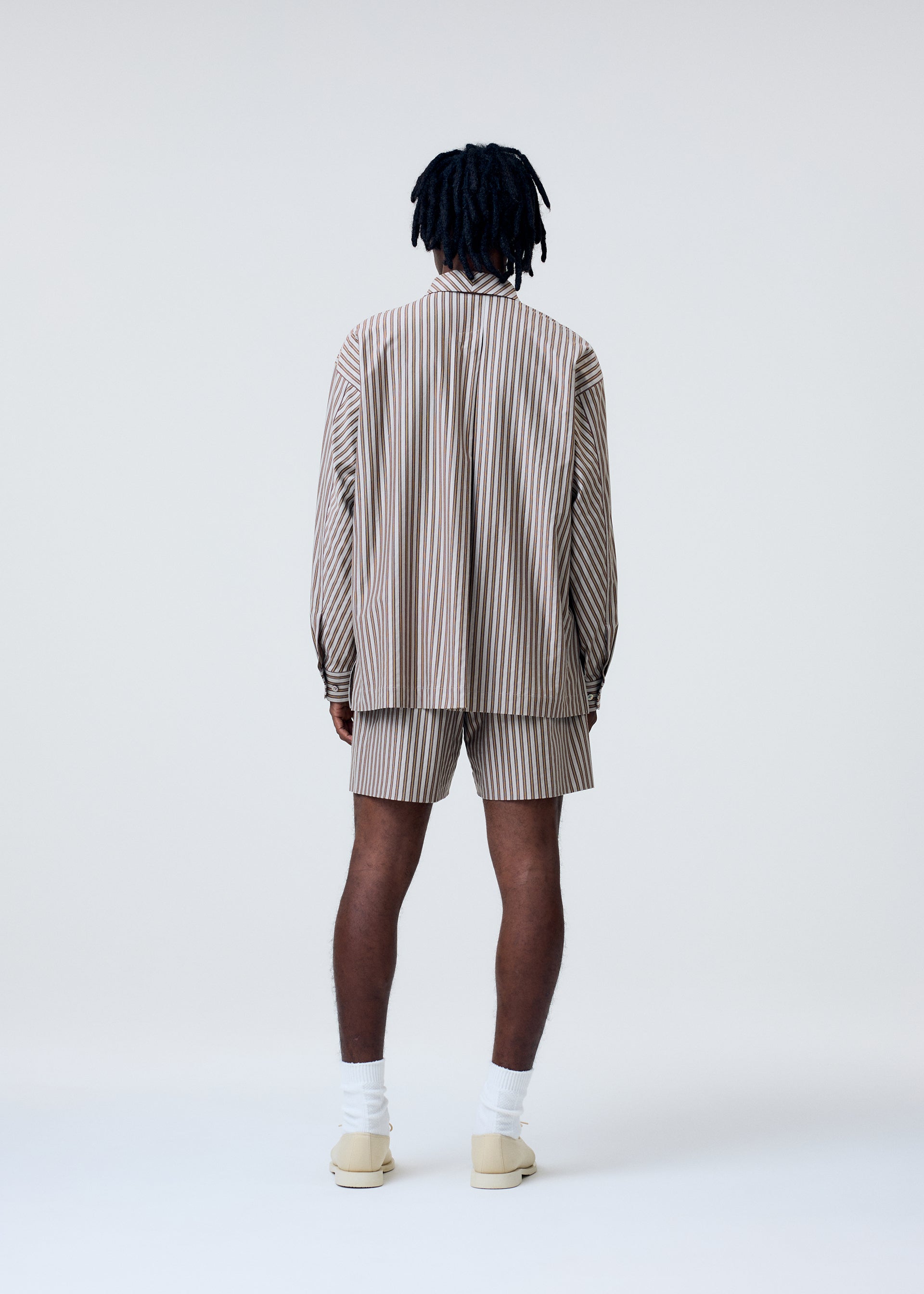 Zoomed view A person with dark, shoulder-length hair faces away, wearing the HOMME PLISSÉ ISSEY MIYAKE STRIPE JERSEY set in beige and brown, paired with white socks and beige shoes, against a plain light background.