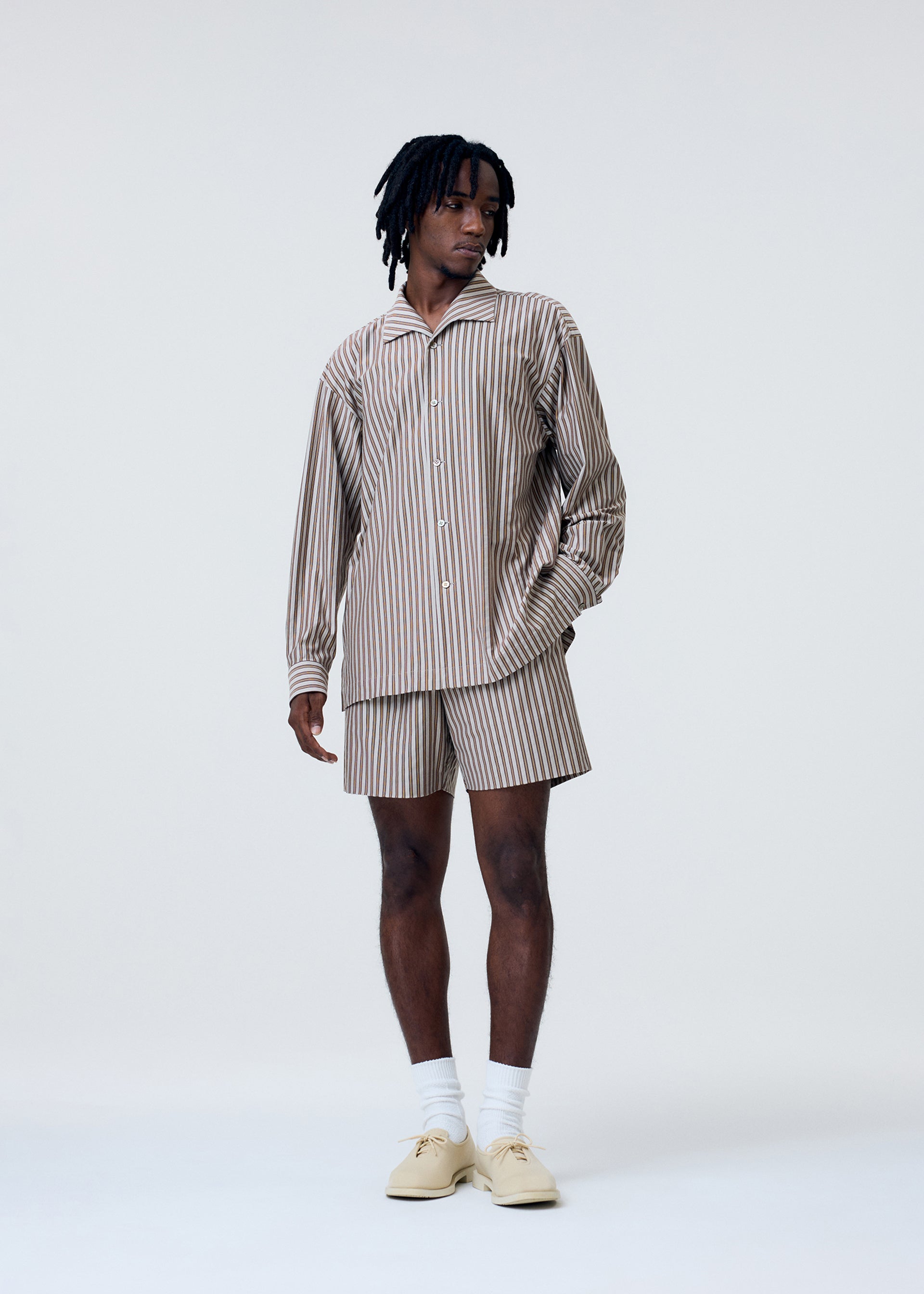 Zoomed view A person stands against a plain light background wearing the STRIPE JERSEY by HOMME PLISSÉ ISSEY MIYAKE—beige and white vertically striped shirt and matching shorts, paired with white socks and cream shoes. They look slightly to the side, one hand in their pocket.