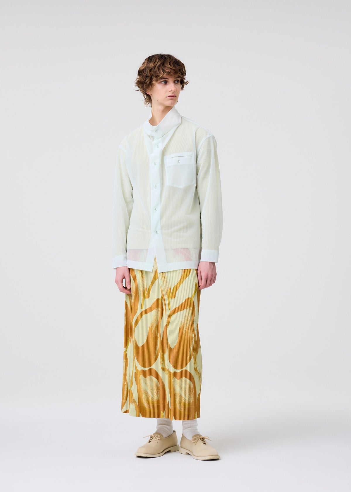 A person with short curly hair stands against a white background, wearing the HOMME PLISSÉ ISSEY MIYAKE FRUITS & VEGETABLES yellow pleated skirt with Italian market prints, styled with a sheer white shirt over a collared top. view 6 of 9