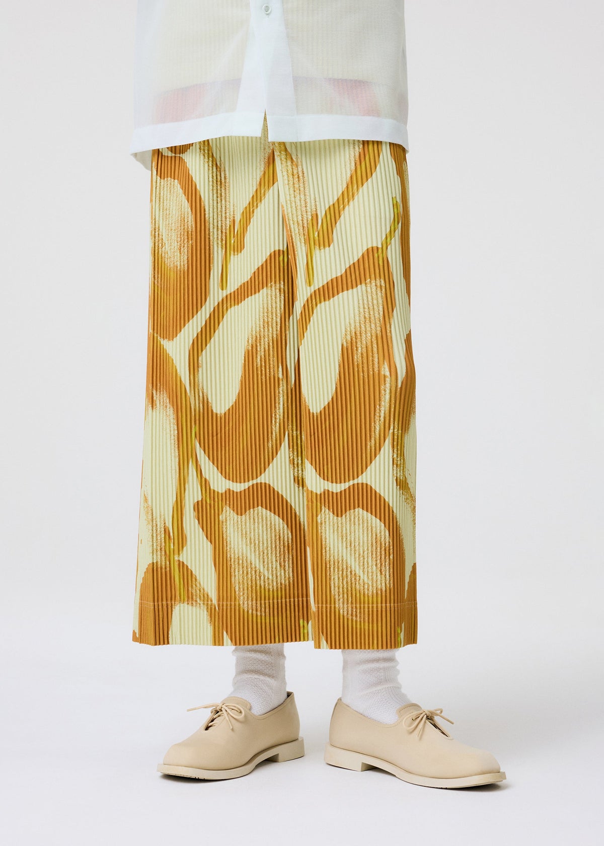 A person stands against a plain white background wearing HOMME PLISSÉ ISSEY MIYAKE’s FRUITS & VEGETABLES Italian market print pleated skirt, a white shirt, white socks, and beige lace-up shoes. view 8 of 9