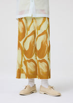 A person stands against a plain white background wearing HOMME PLISSÉ ISSEY MIYAKE’s FRUITS & VEGETABLES Italian market print pleated skirt, a white shirt, white socks, and beige lace-up shoes.