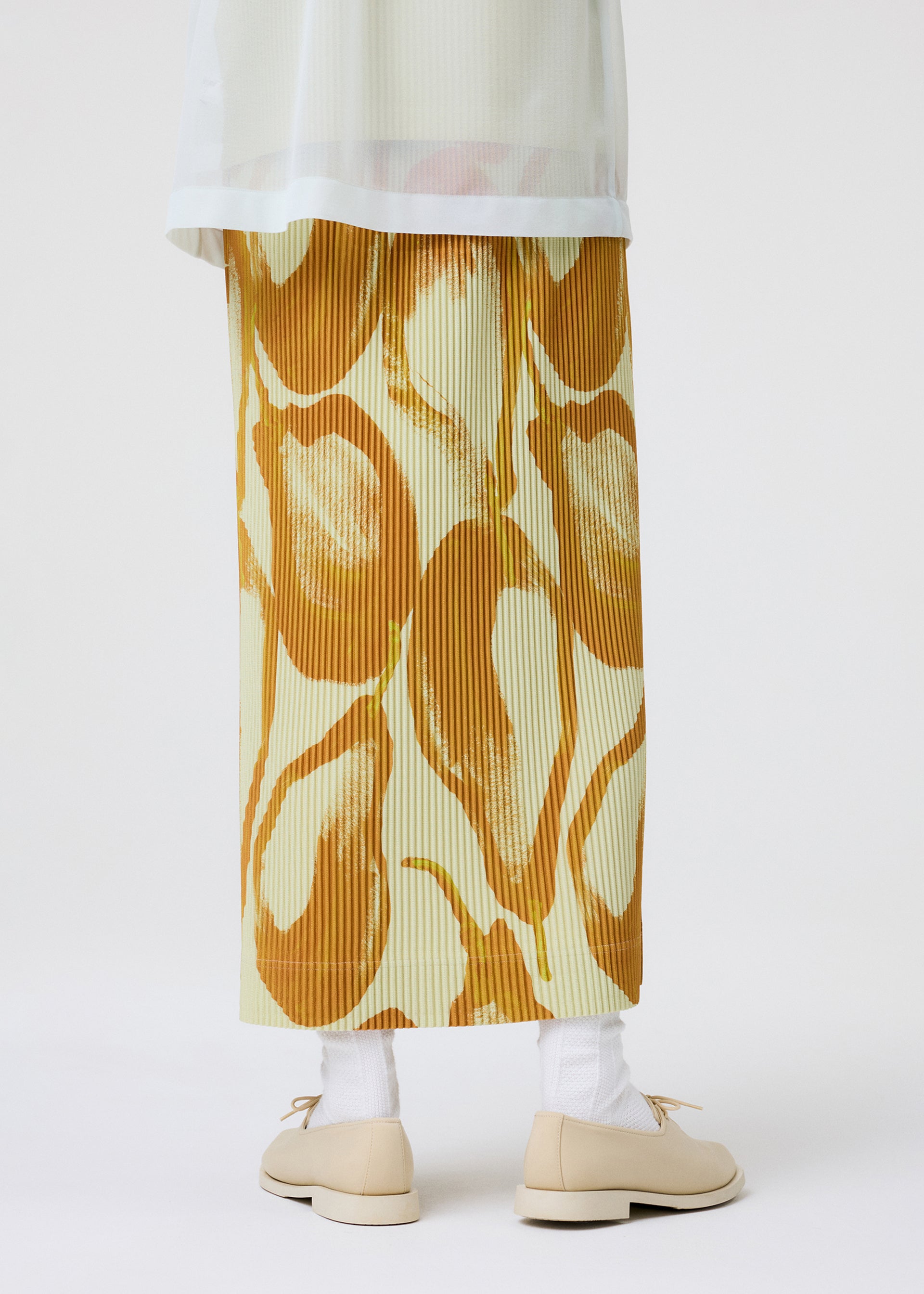 Zoomed view A person stands against a white background wearing the HOMME PLISSÉ ISSEY MIYAKE FRUITS & VEGETABLES long, pleated skirt with a colorful food motif, paired with white socks, beige shoes, and a sheer light top.