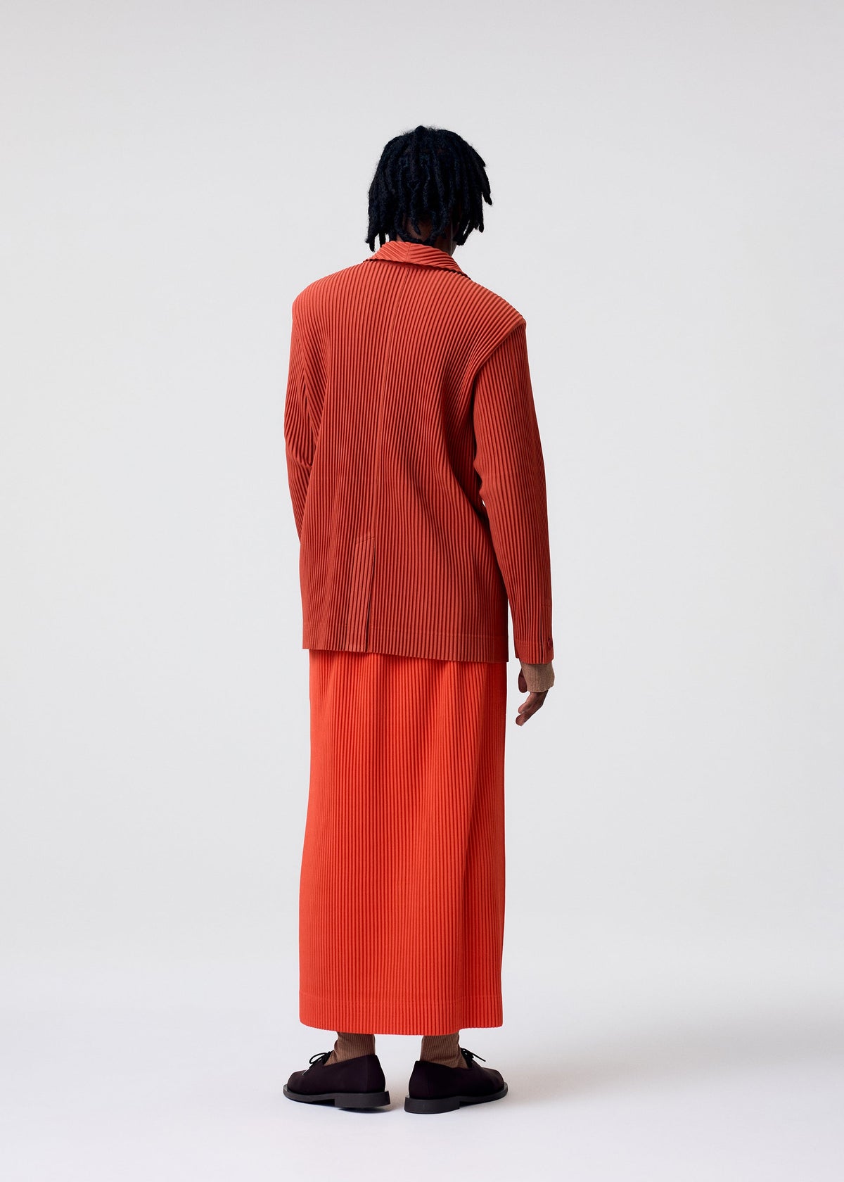 A person with dark, shoulder-length hair stands facing away, wearing HOMME PLISSÉ ISSEY MIYAKE COLORFUL PLEATS BOTTOMS in orange with a matching top and dark sandals, set against a plain light background. view 8 of 8
