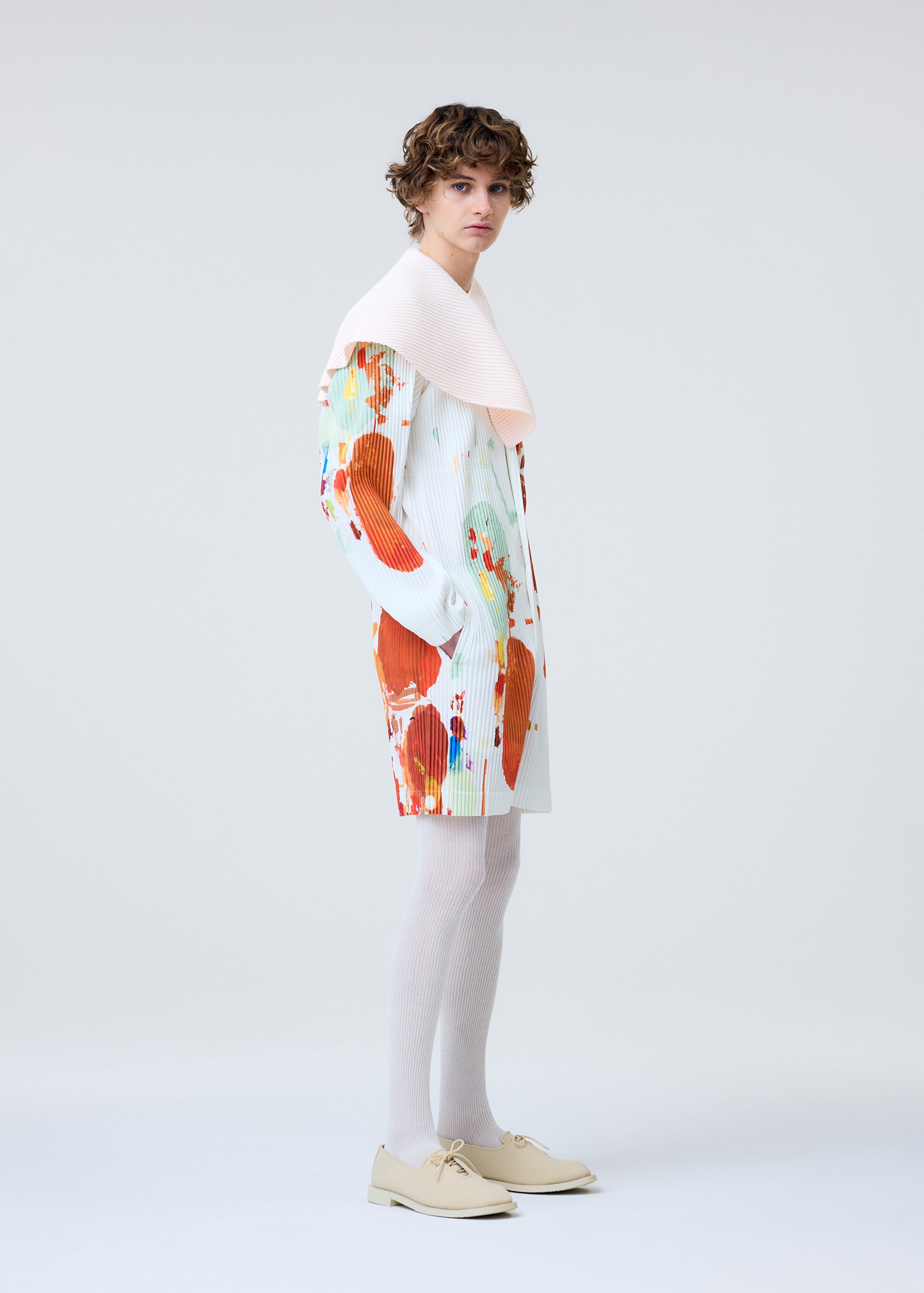 Zoomed view A person with short curly hair wears the PALETTE coat by HOMME PLISSÉ ISSEY MIYAKE, featuring colorful abstract patterns and a pale pink collar, paired with white tights and beige shoes against a plain light background.