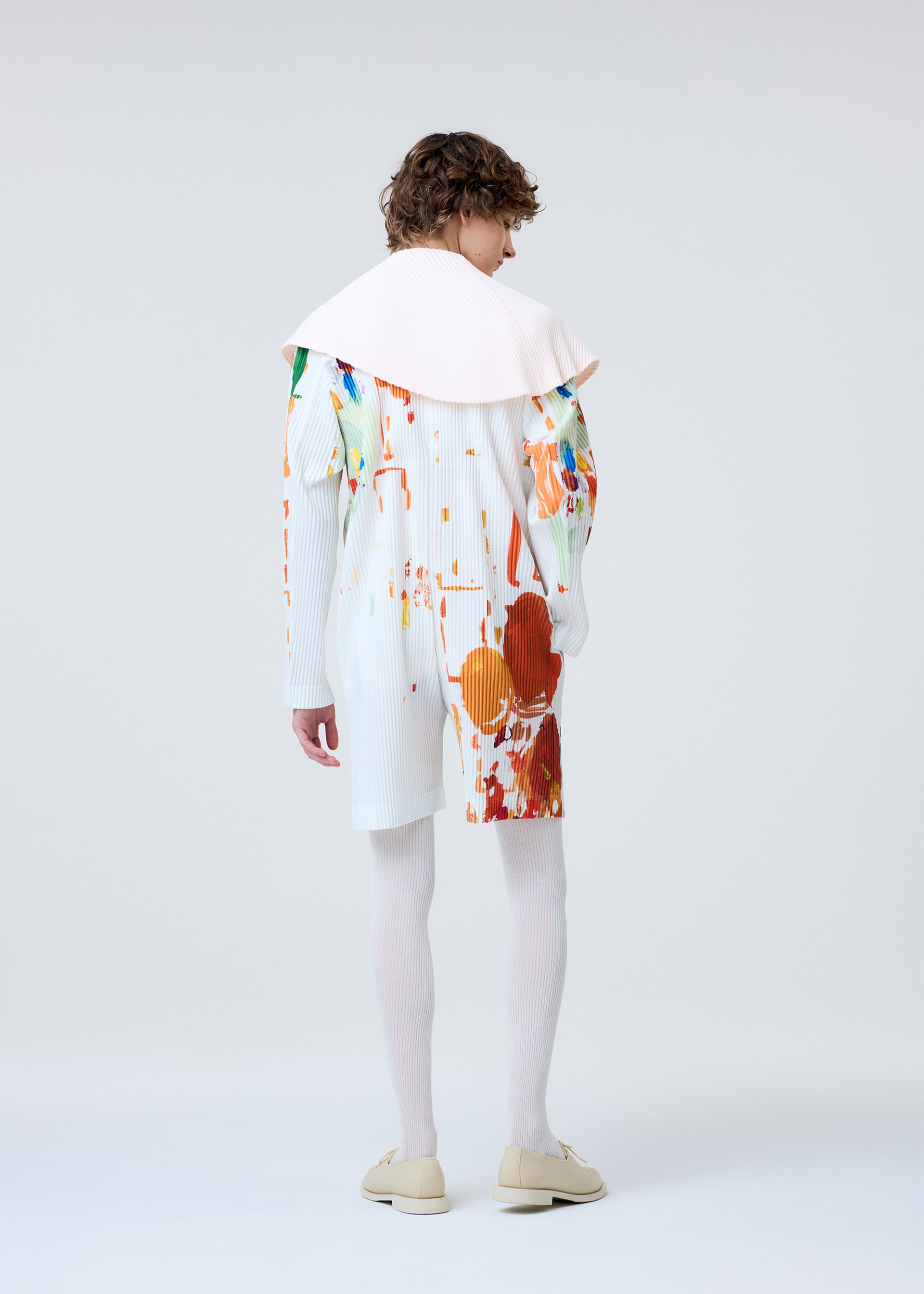 Zoomed view A person with curly hair stands facing away, wearing HOMME PLISSÉ ISSEY MIYAKE's PALETTE: a white outfit splattered with colorful paint, white knee-high socks, beige shoes, and a large pale pink collar against a plain light background.