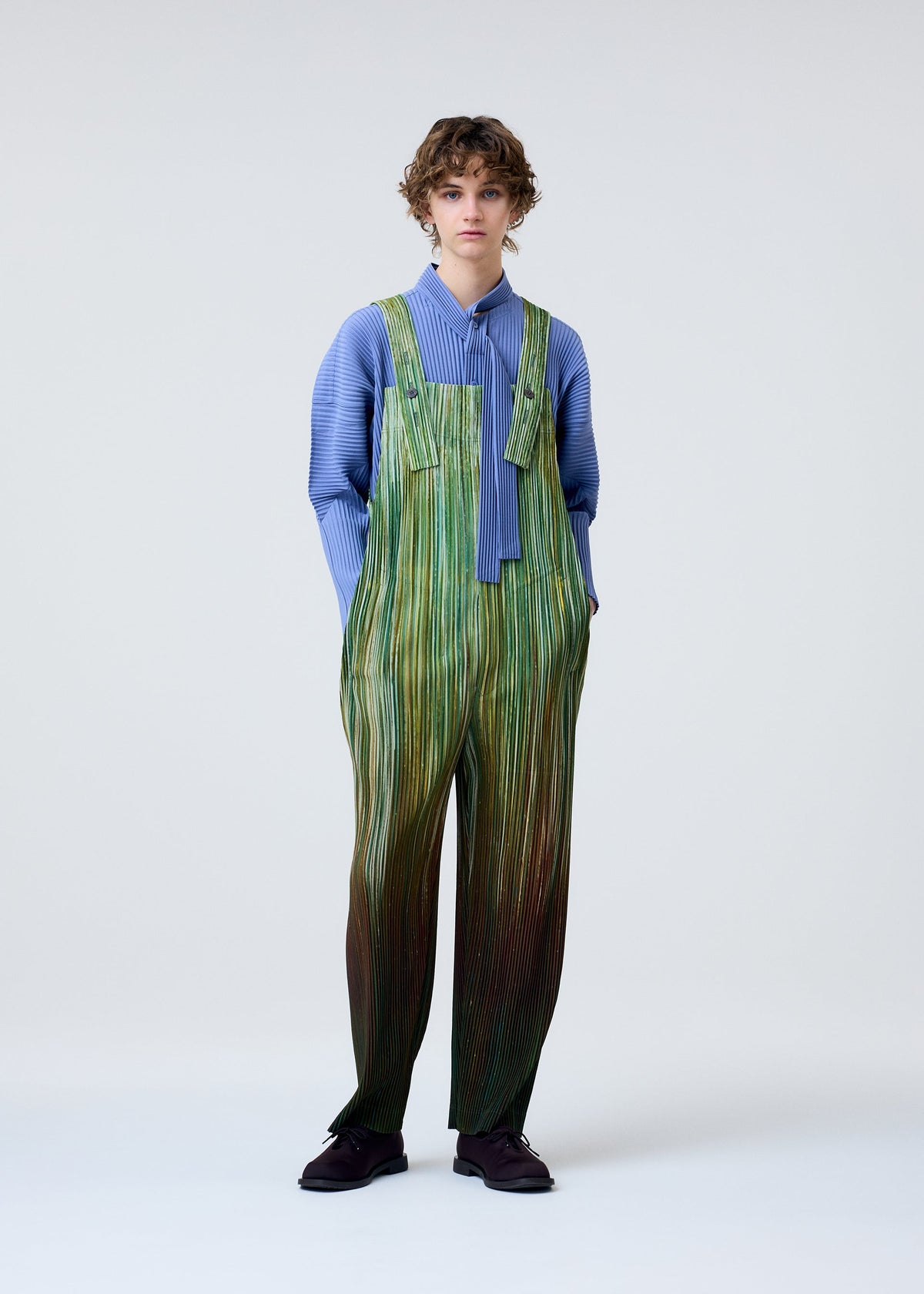A person with short curly hair models HOMME PLISSÉ ISSEY MIYAKE's PAINT BRUSH CLOSE-UP: a blue long-sleeve top with tie detail layered under green-striped overalls, standing against a plain light background, hands in pockets. view 6 of 7
