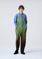 A person with short curly hair models HOMME PLISSÉ ISSEY MIYAKE's PAINT BRUSH CLOSE-UP: a blue long-sleeve top with tie detail layered under green-striped overalls, standing against a plain light background, hands in pockets.