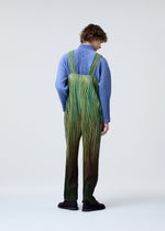 A person with curly hair, wearing HOMME PLISSÉ ISSEY MIYAKE's PAINT BRUSH CLOSE-UP: a ribbed blue shirt and a loose, green-and-brown striped jumpsuit with layered patterns, stands against a plain light background.