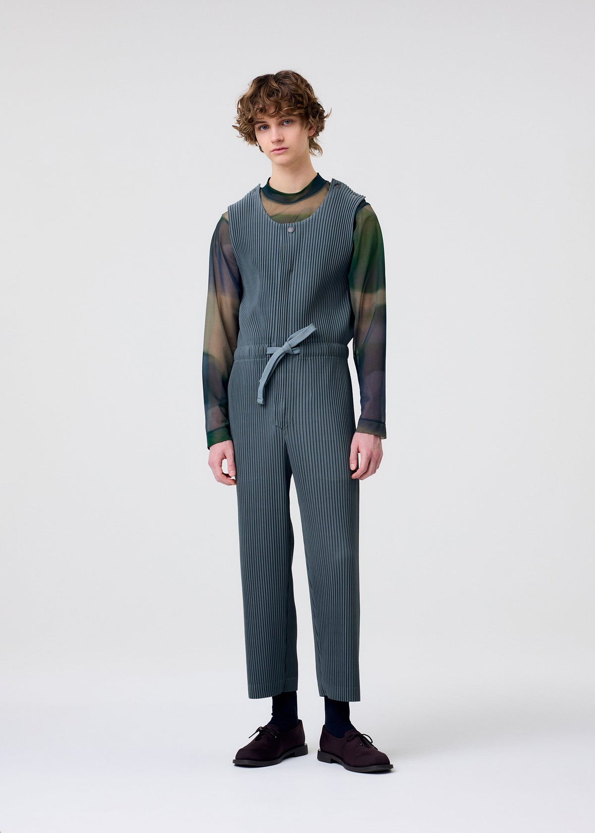 A person with curly hair stands against a white background, wearing HOMME PLISSÉ ISSEY MIYAKE PLEATS BOTTOMS 2 paired with a sheer green and blue long-sleeve shirt, dark socks, and brown shoes. view 6 of 7