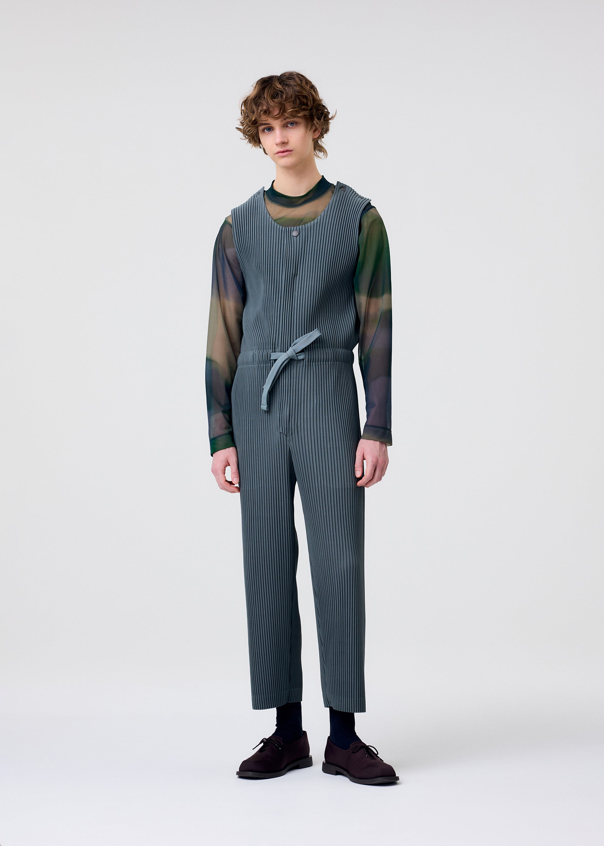 Zoomed view A person with curly hair stands against a white background, wearing HOMME PLISSÉ ISSEY MIYAKE PLEATS BOTTOMS 2 paired with a sheer green and blue long-sleeve shirt, dark socks, and brown shoes.