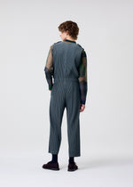Facing away, a person with curly hair wears HOMME PLISSÉ ISSEY MIYAKE's PLEATS BOTTOMS 2 over a sheer dark long-sleeve shirt, matched with dark socks and brown shoes, set against a light gray background.