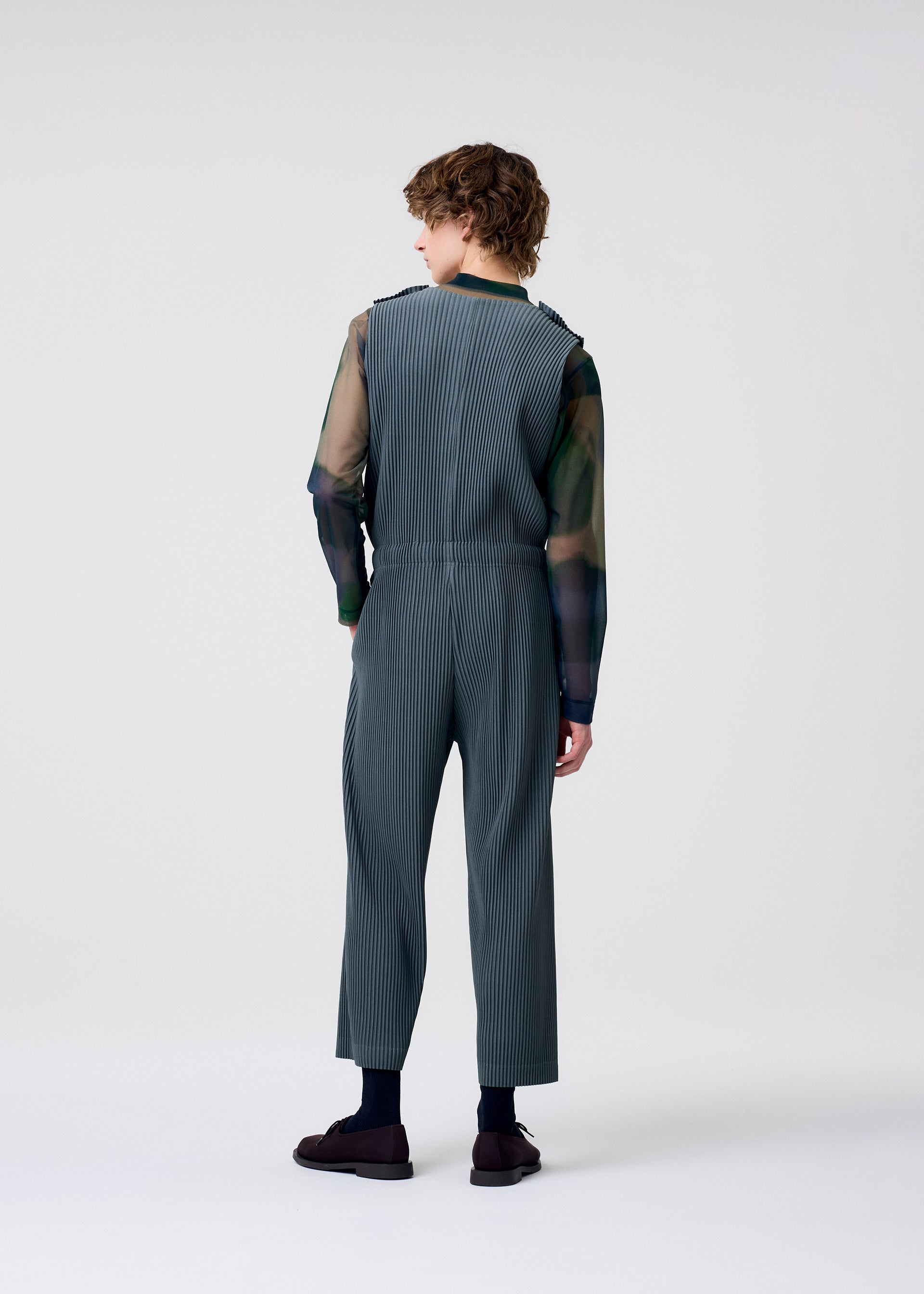 Zoomed view Facing away, a person with curly hair wears HOMME PLISSÉ ISSEY MIYAKE's PLEATS BOTTOMS 2 over a sheer dark long-sleeve shirt, matched with dark socks and brown shoes, set against a light gray background.