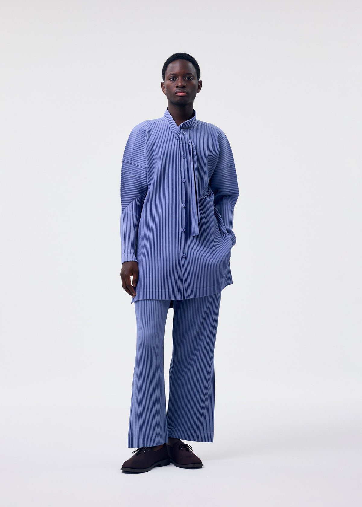 A person stands against a plain light background, wearing the HOMME PLISSÉ ISSEY MIYAKE MONTHLY COLORS : FEBRUARY set—long, loose-fitting blue pinstriped shirt and matching wide-leg trousers—with dark shoes, one hand in their pocket and a neutral expression. view 5 of 6