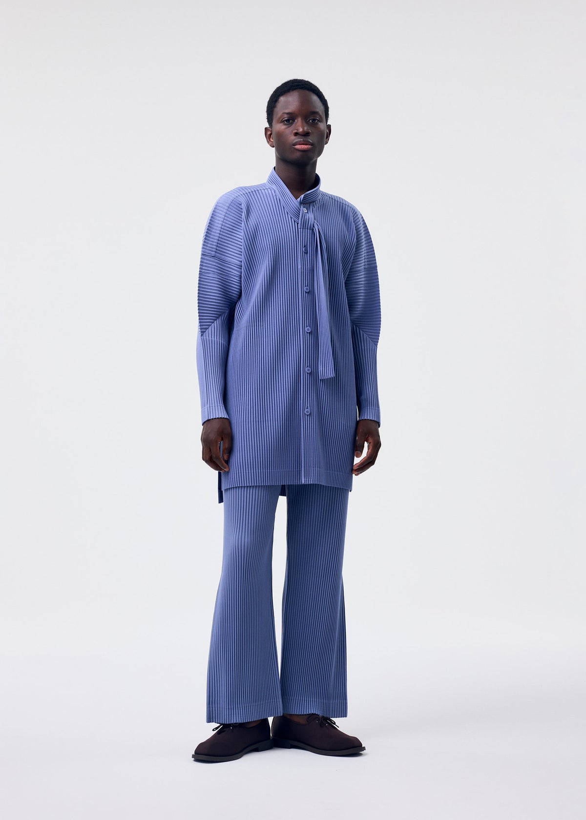 A person with short hair and dark shoes stands against a plain background, wearing the HOMME PLISSÉ ISSEY MIYAKE MONTHLY COLORS : FEBRUARY set—blue pinstriped long-sleeved shirt with flared sleeves and matching flared pants. view 6 of 6