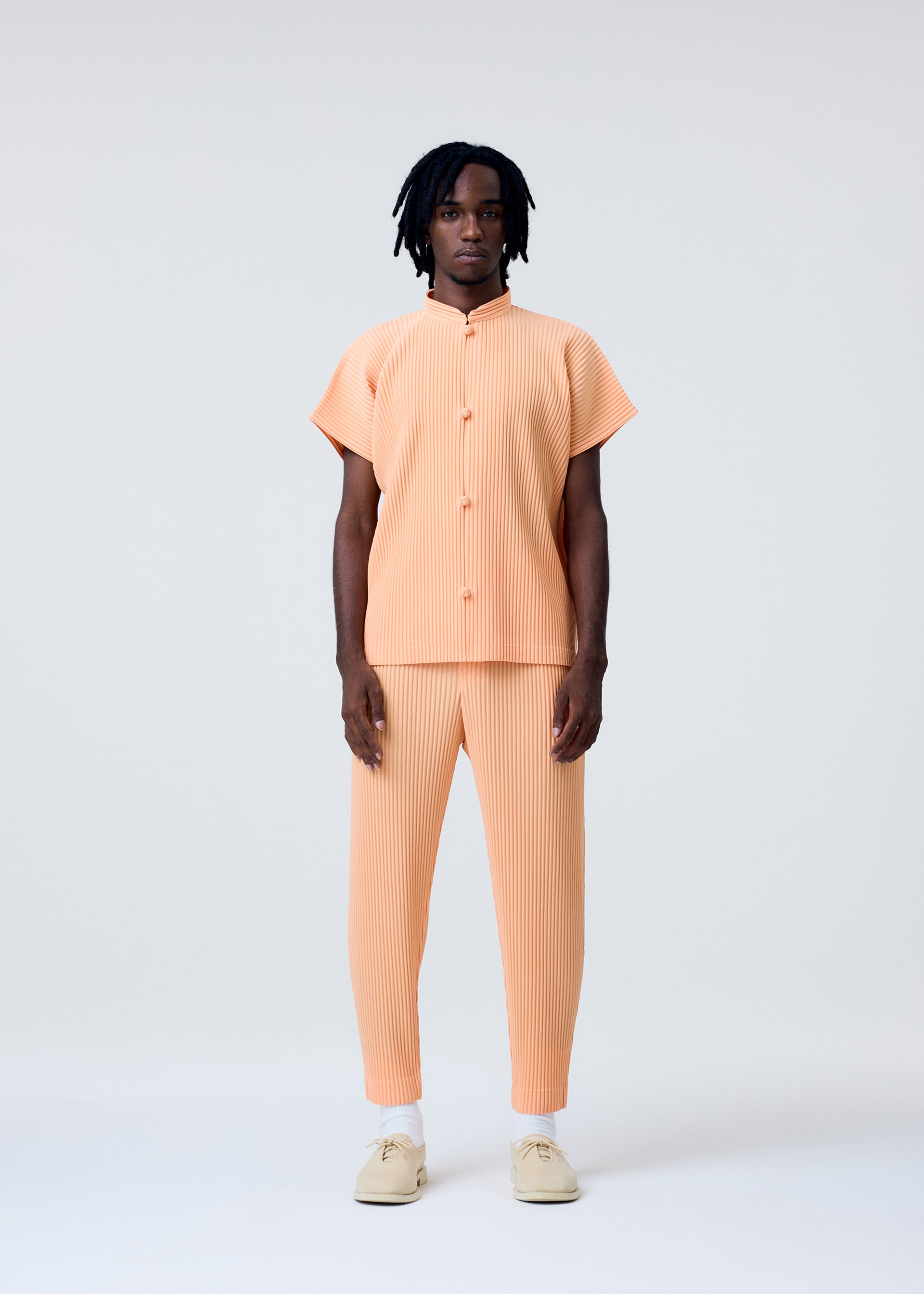 Zoomed view A person with short dreadlocks faces forward against a plain background, wearing HOMME PLISSÉ ISSEY MIYAKE’s MONTHLY COLORS : APRIL—a matching peach-toned, pleated, short-sleeve shirt and pants set—paired with light-colored shoes.