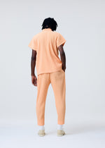 A person with dark hair faces away, wearing HOMME PLISSÉ ISSEY MIYAKE's MONTHLY COLORS: APRIL—a peach-toned ribbed quick-drying shirt and matching pants—with white socks and beige slip-ons against a light background.