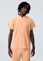 A person with medium-length dreadlocks models the MONTHLY COLORS : APRIL set by HOMME PLISSÉ ISSEY MIYAKE—a light orange pleated short-sleeve top with Chinese-style buttons and matching pants—against a plain, light background.