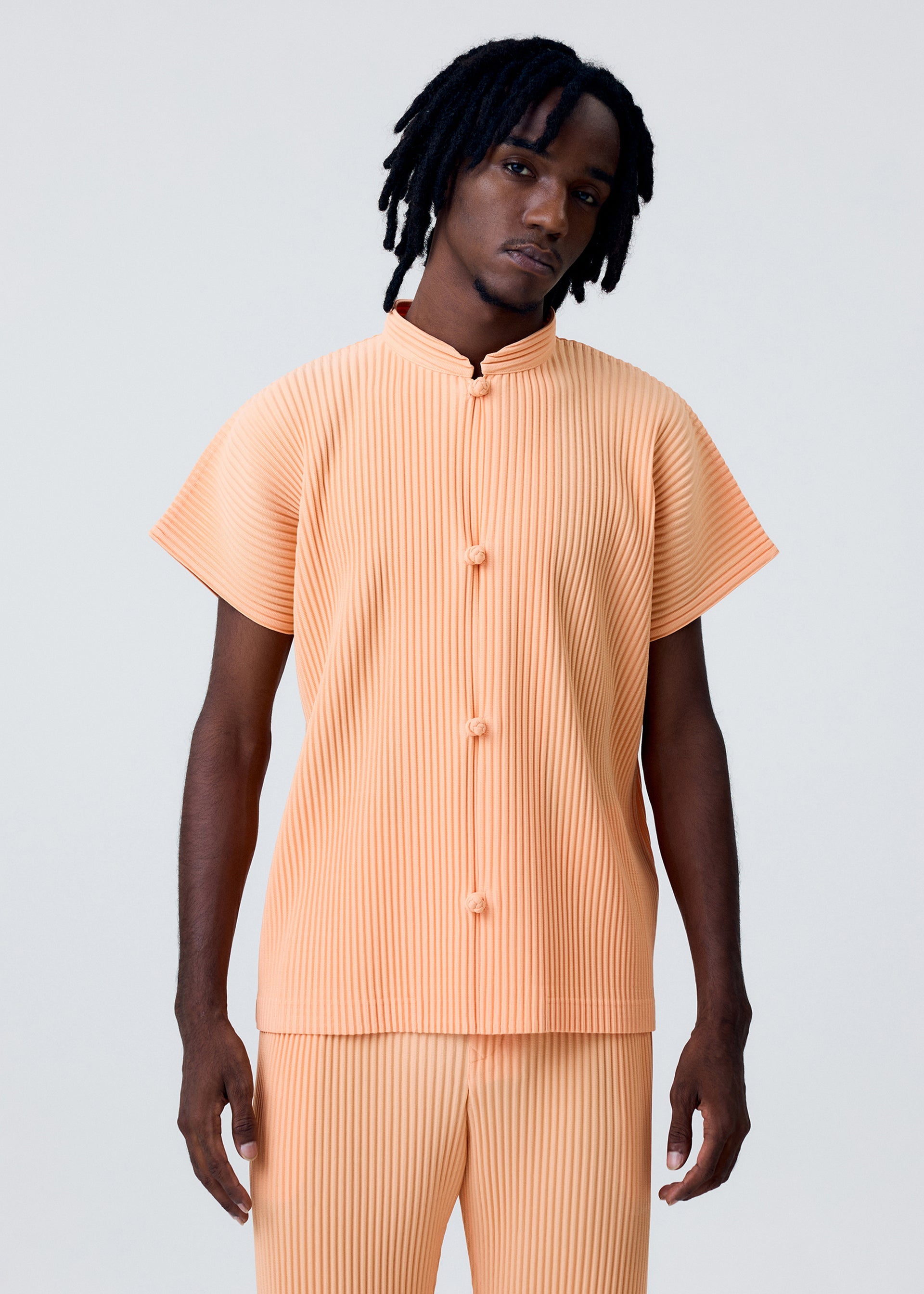 Zoomed view A person with medium-length dreadlocks models the MONTHLY COLORS : APRIL set by HOMME PLISSÉ ISSEY MIYAKE—a light orange pleated short-sleeve top with Chinese-style buttons and matching pants—against a plain, light background.